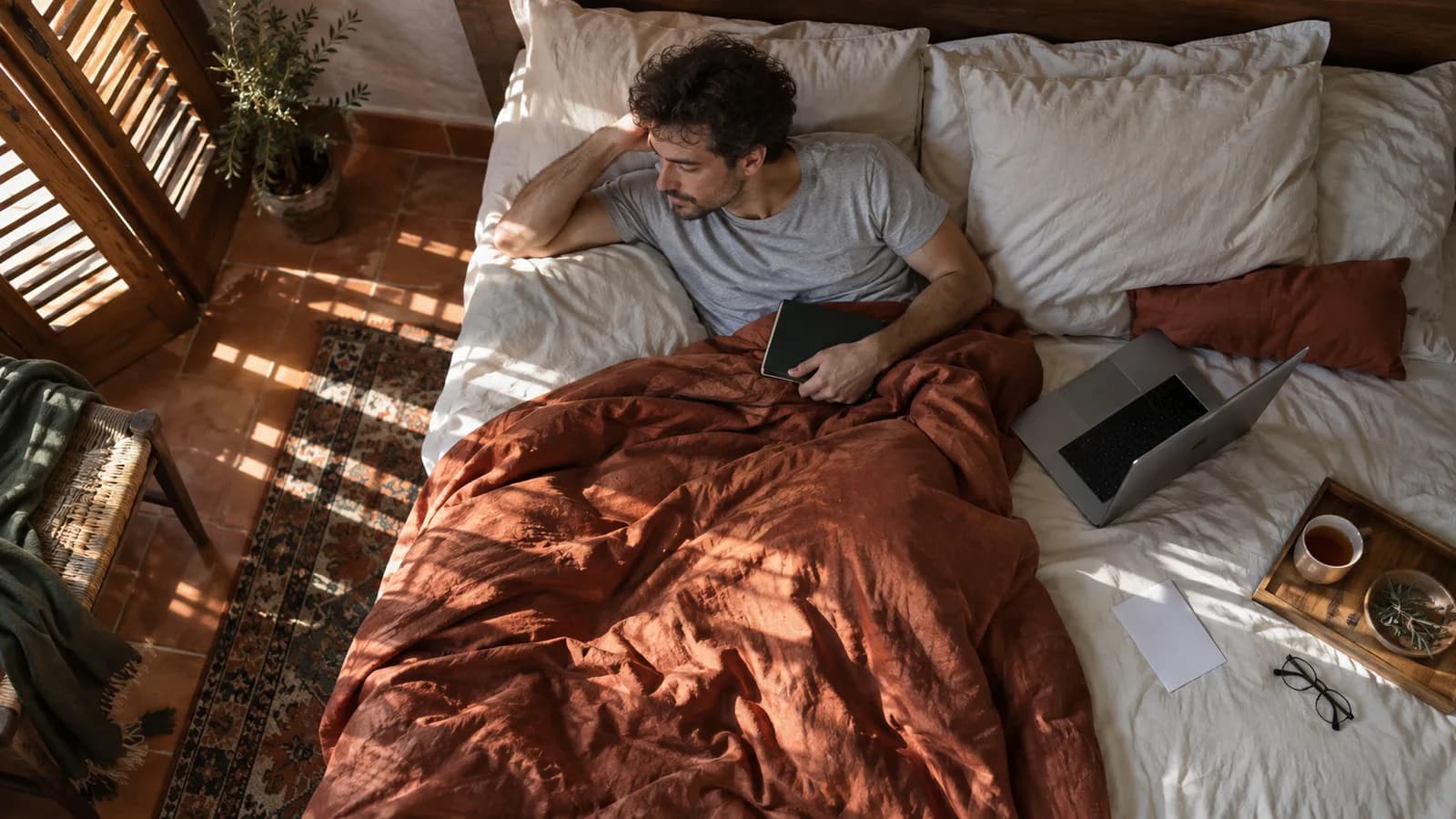 Hombre de baja por enfermedad descansando en la cama con un portátil y una infusión, visto desde arriba.